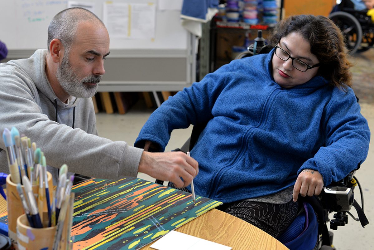 A young woman in a wheelchair gives instructions to a man sitting next to her. He is holding a paintbrush to a canvas board, which has an in-progress acrylic painting on it.