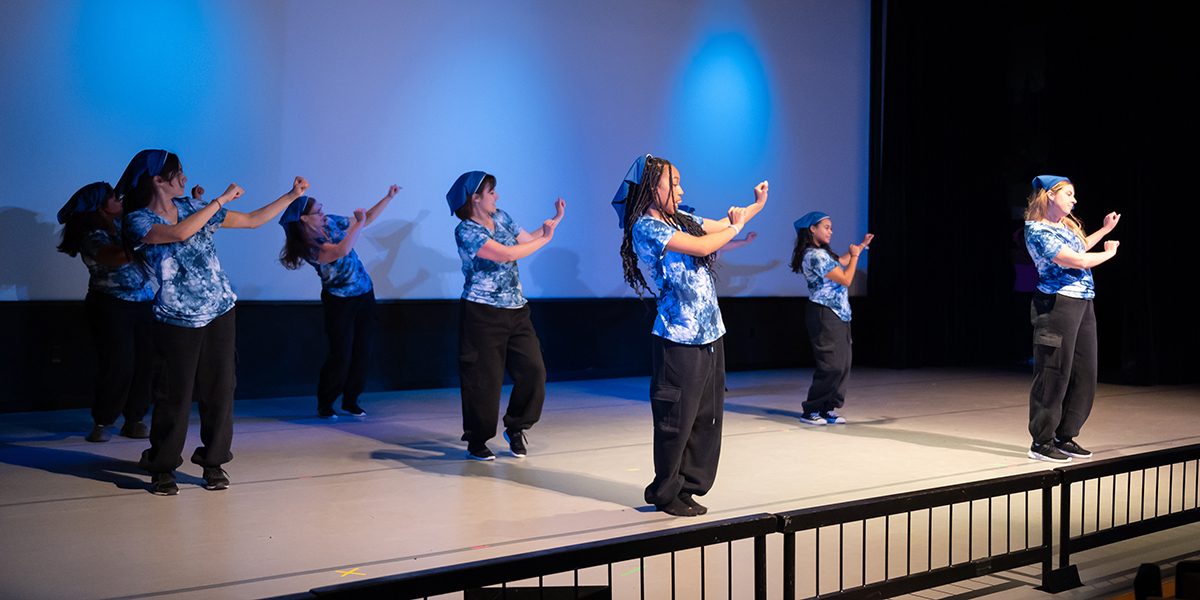 Seven able-bodied dancers onstage, wearing blue tie-dye shirts, blue bandanas on their heads, and black cargo pants. They are standing in a grid, dancing in a hip-hop style.