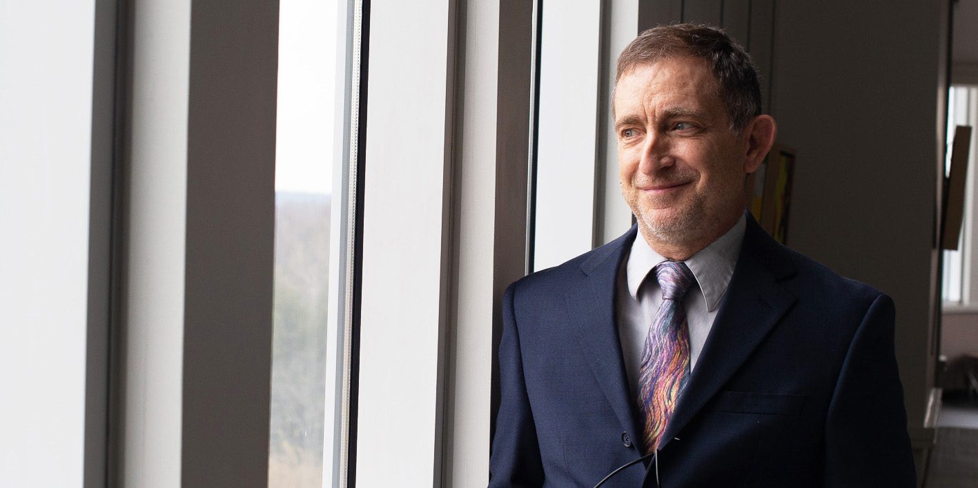 A man wearing a suit and an Arts Access silk tie, standing next to a series of tall, thin windows.