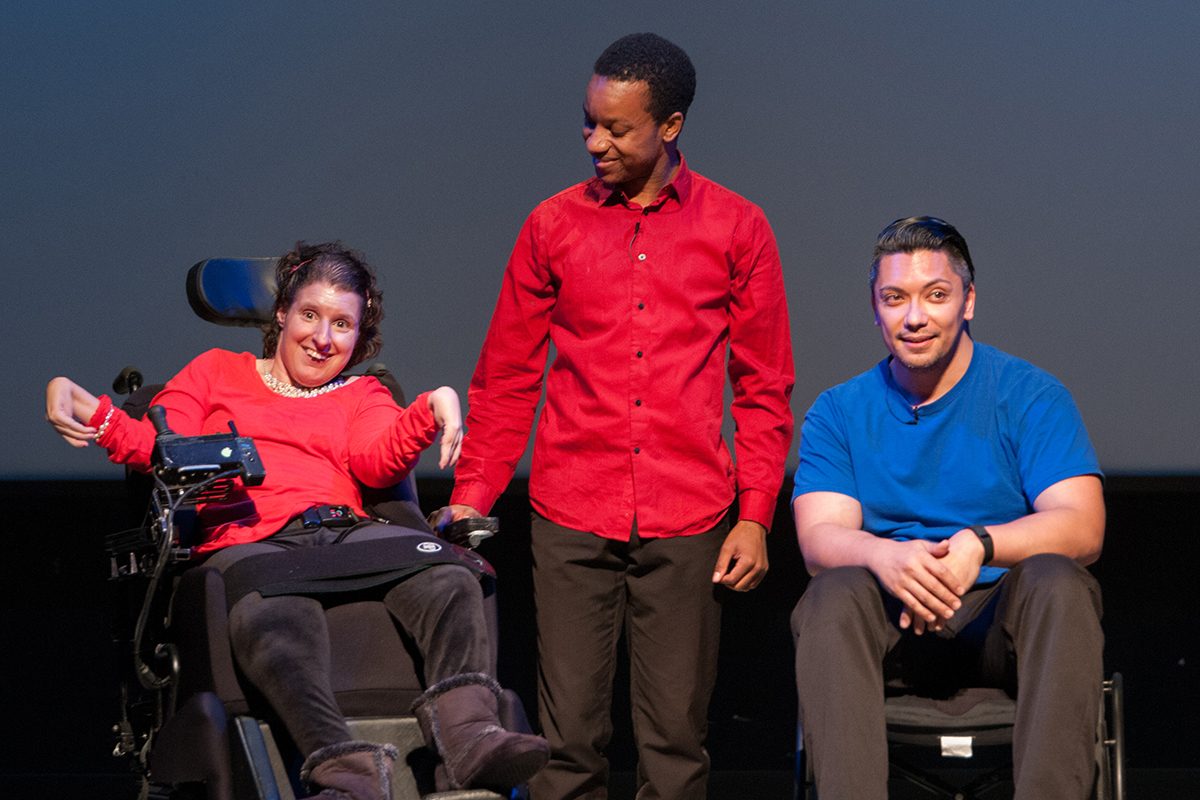 Three actors are arranged onstage in a line, side-by-side. On the left is a young woman in a power wheelchair, in the center is a man standing, on the right is another man in a manual wheelchair.