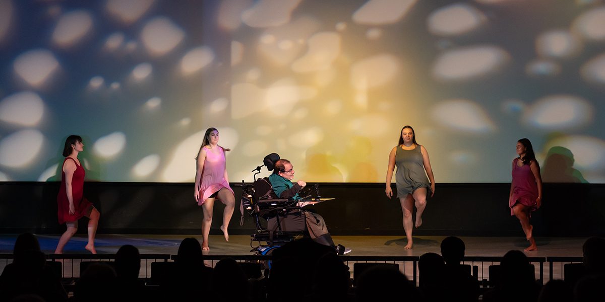 Four female able-bodied dancers and one male dancer using a power wheelchair performing onstage at Full Circle 2023