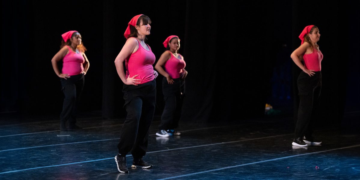 Four female dancers wearing pink tank tops and bandanas on their heads with black pants, on a stage.