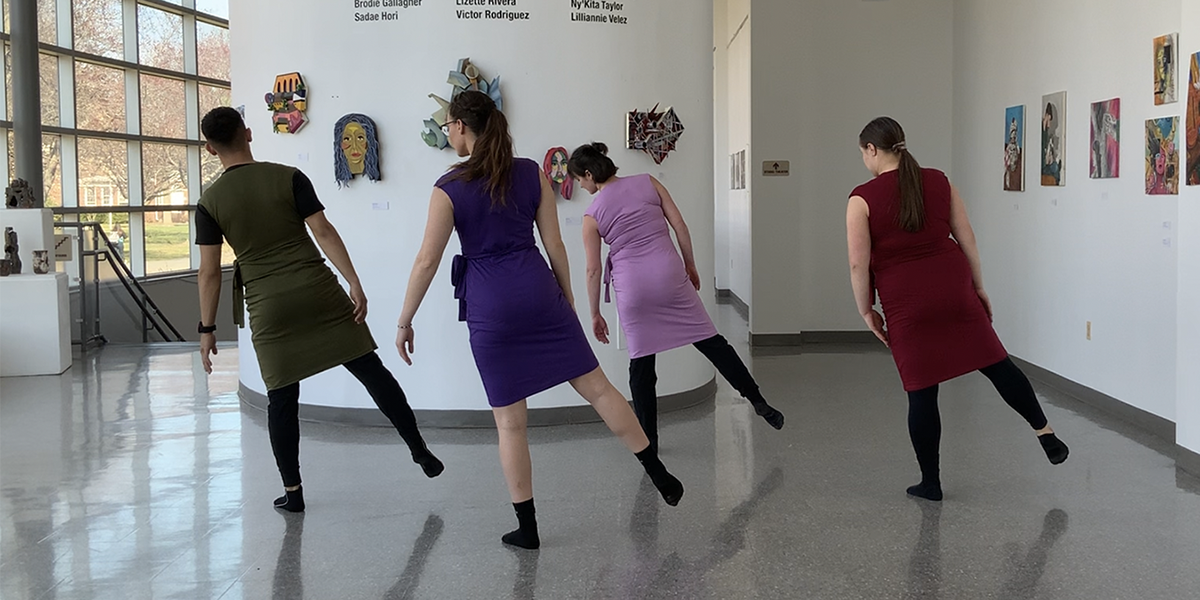 Four dancers in different colored dresses, backs facing the viewer, in an art gallery.