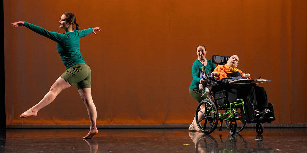 One man in a wheelchair and two able-bodied women dancing on a stage with an orange backdrop