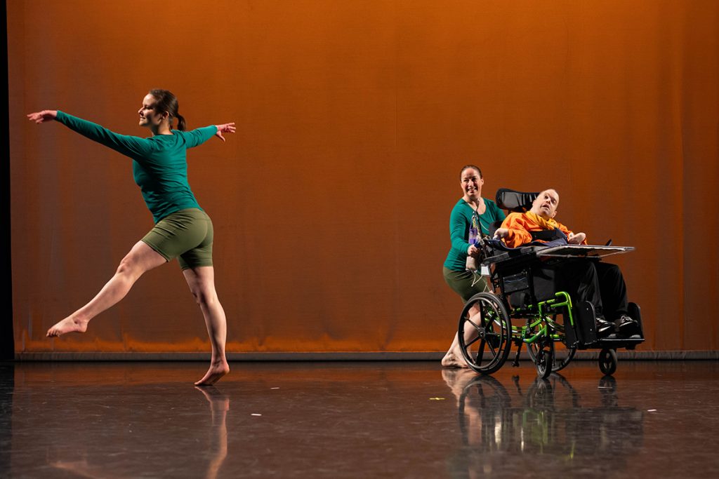 One man in a wheelchair and two able-bodied women dancing on a stage with an orange backdrop