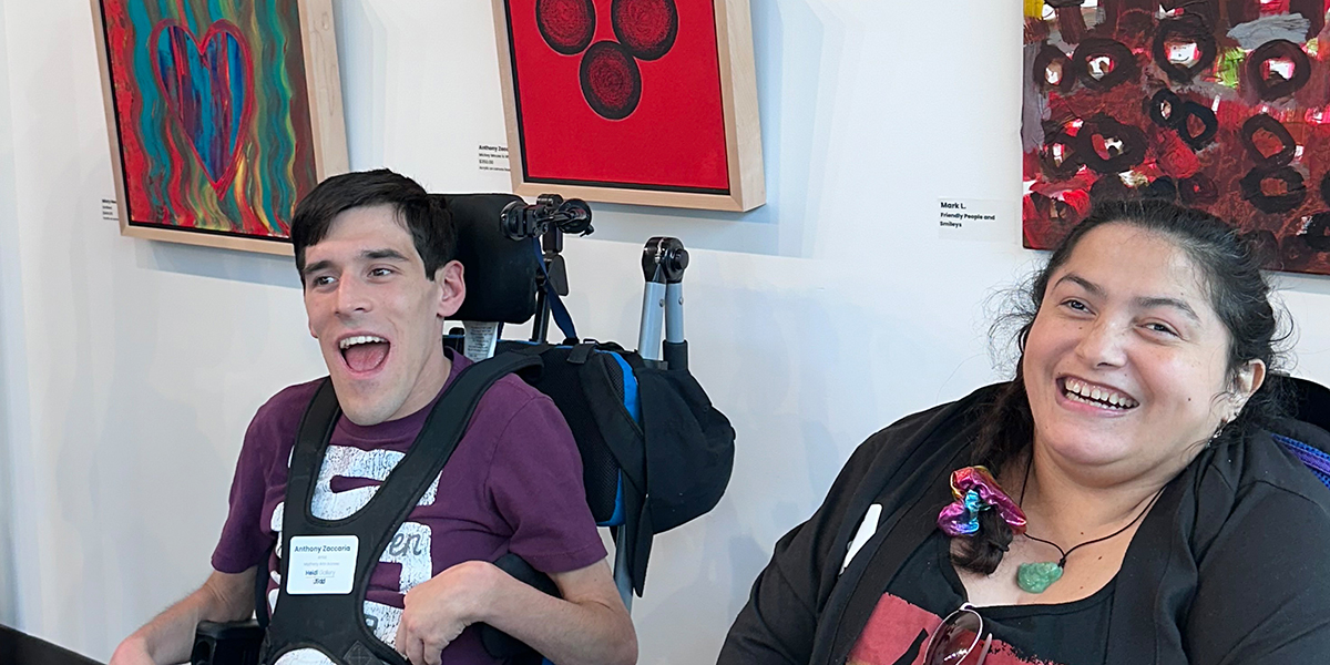 A young man and woman in wheelchairs, sitting in front of a gallery wall full of abstract paintings.