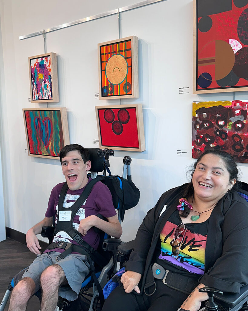 A young man and woman in wheelchairs, sitting in front of a gallery wall full of abstract paintings.