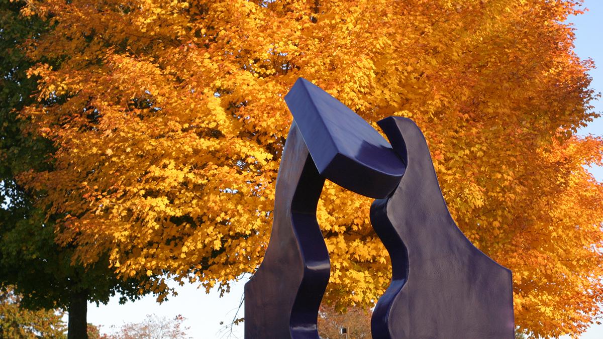 large blue abstract sculpture outside in front of a tree full of autumn orange leaves