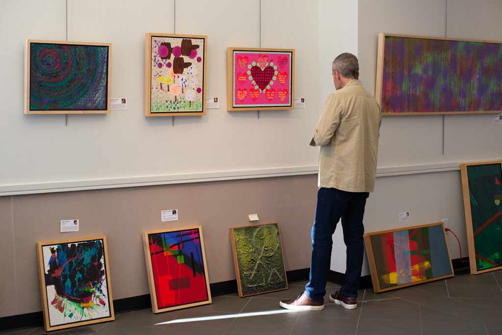 A man looking at abstract acrylic paintings hanging on a gallery wall, and more paintings leaning against the wall on the floor.