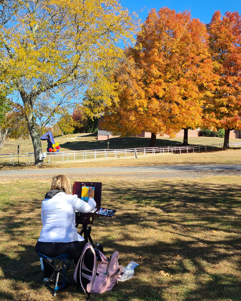 Woman painting in plein air on an autumn day, looking at trees with bright orange leaves