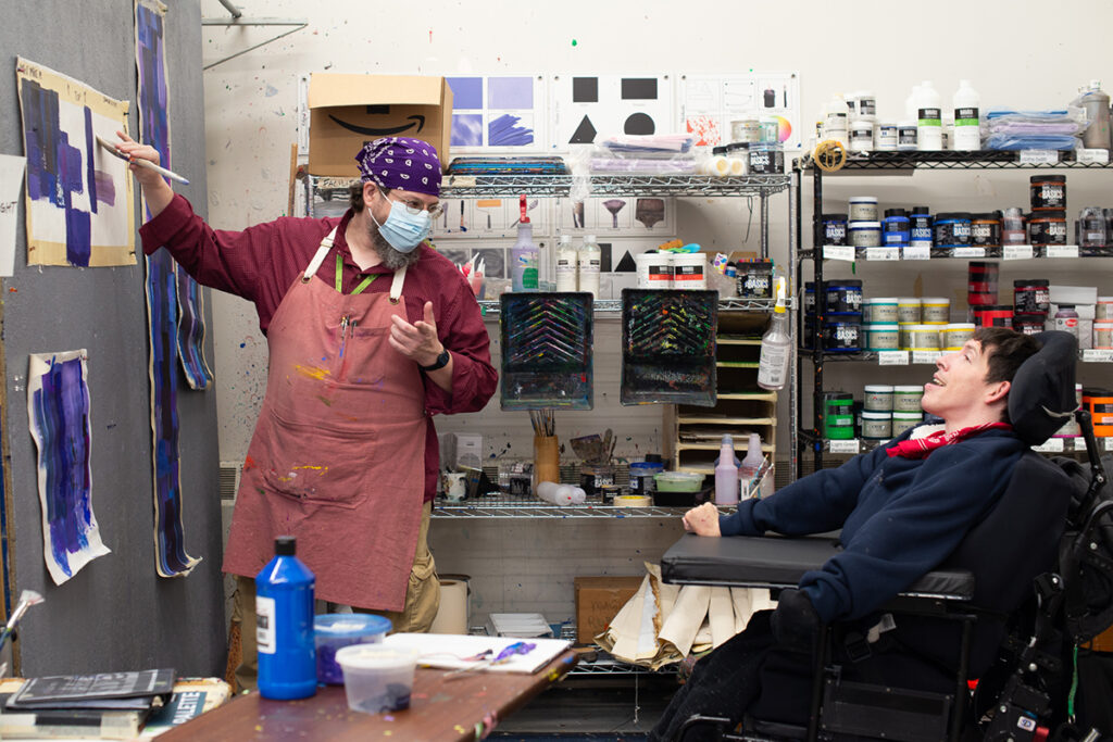 Man in a wheelchair and an able-bodied man in a painting studio, working together on an in-progress abstract acrylic painting