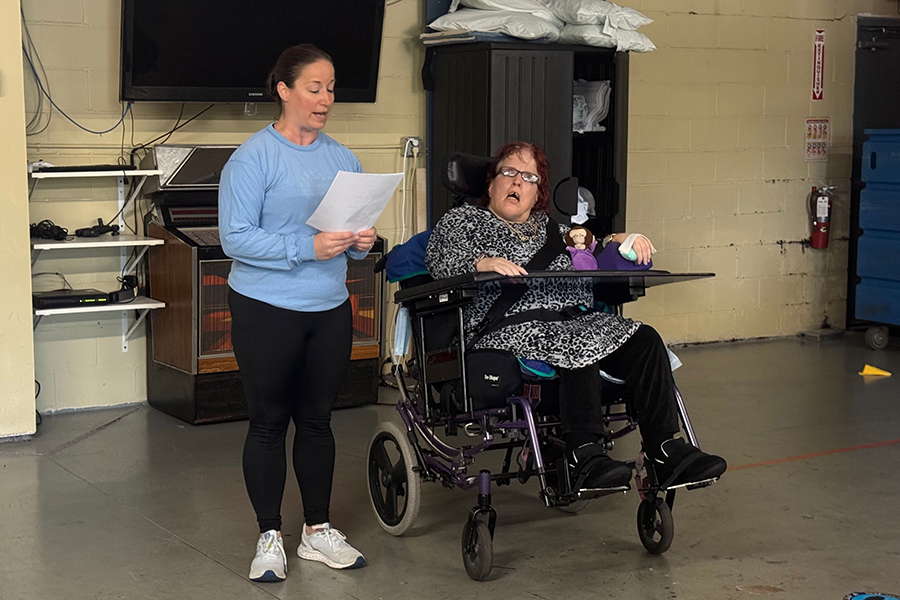 An able-bodied woman reading from a paper held in her hand, standing next to a woman in a wheelchair.