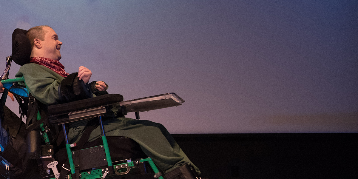 A man in a wheelchair, in profile, on a stage with a soft, blue and purple gradient on the backdrop behind him.