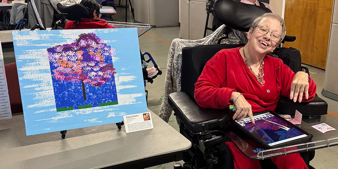 A woman in a wheelchair sitting next to an abstract acrylic painting on a table easel on a folding table.