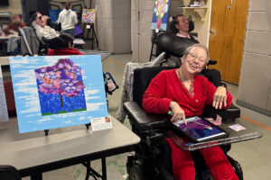 A woman in a wheelchair sitting next to an abstract acrylic painting on a table easel on a folding table.