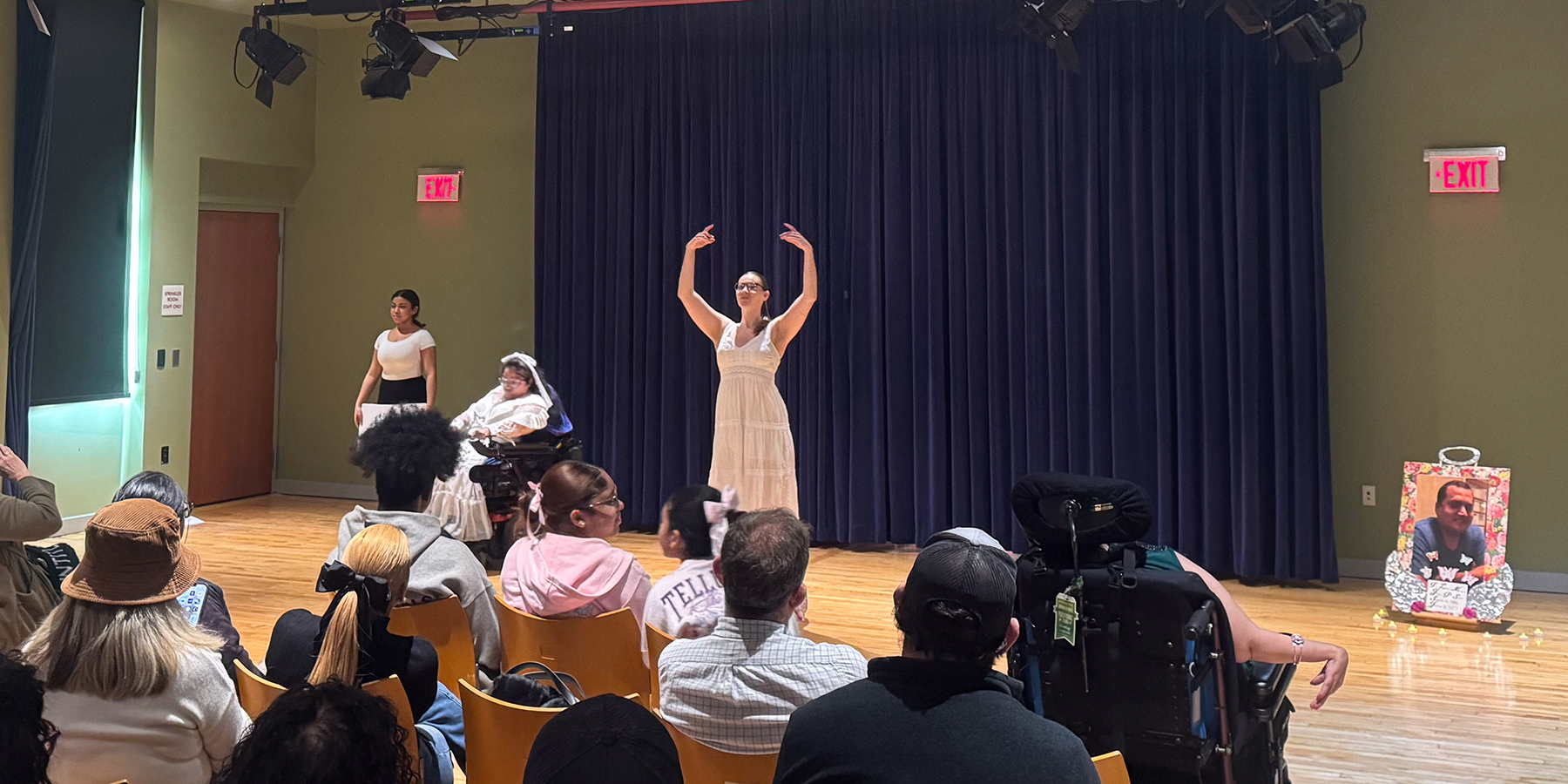 Two able-bodied dancers and one dancer in a wheelchair, wearing white, performing in a black box theater. A large memorial photo sits off to the right side of the performance space.