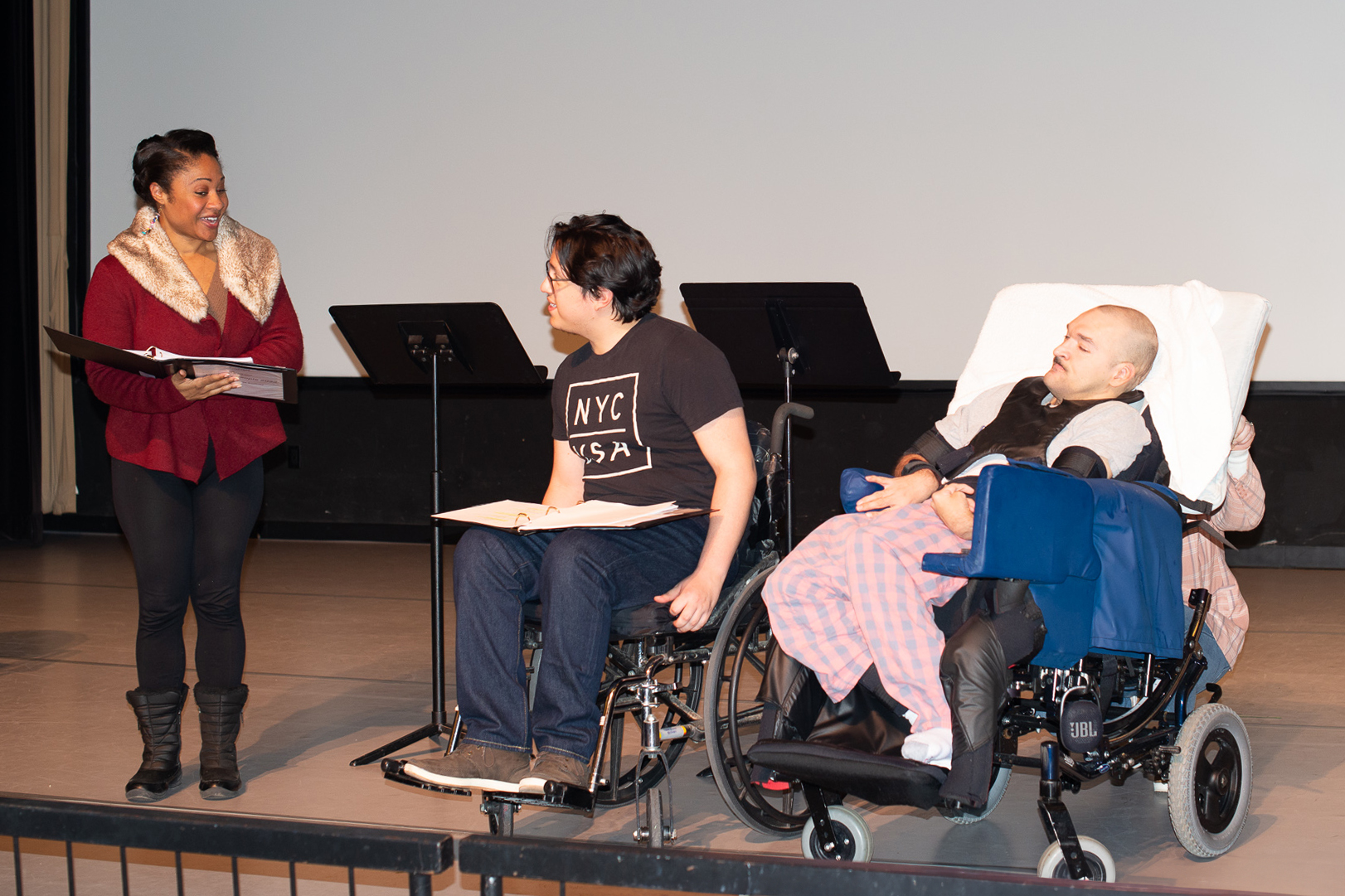 Three actors, one standing and two sitting in wheelchairs, on a stage with scripts in front of them.