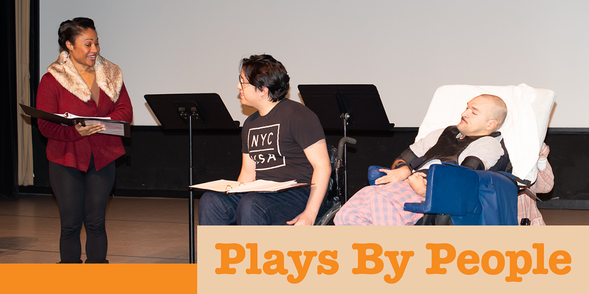 Three actors, one standing and two sitting in wheelchairs, on a stage with scripts in front of them.