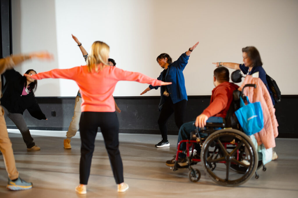 High school students standing in a circle on the stage at Arts Access, practicing creative movement with staff.