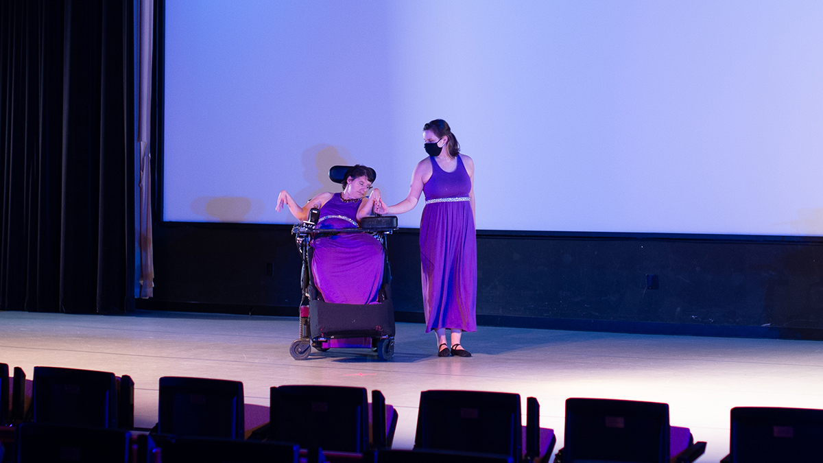 Two women wearing purple dresses, one in a wheelchair and one able-bodied, dancing on a stage.