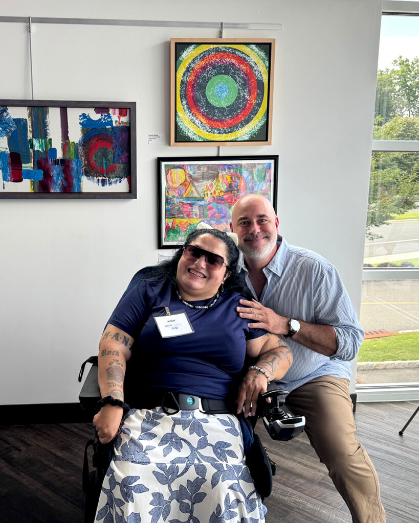A woman in a wheelchair and an able-bodied man smile as they pose in front of abstract art hanging on the gallery walls at the Summer Symbiosis II exhibit.