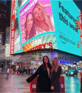 Hallie Stephens Drake and Katie Krause Mork in front of a billboard for their podcast "Between Us Moms."