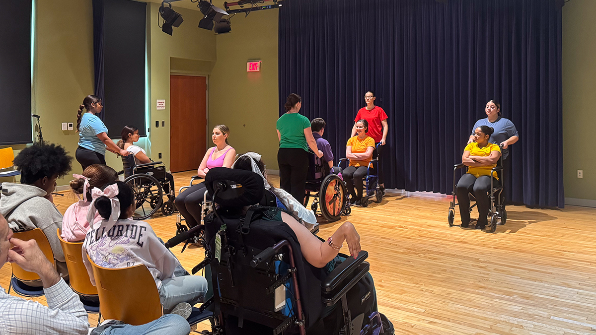 Four able-bodied dancers and five dancers in a wheelchairs, wearing different brightly colored tops, performing in a black box theater.