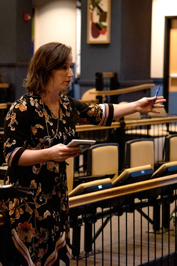An able-bodied woman standing in an empty theatre. She holds a notepad and pen.