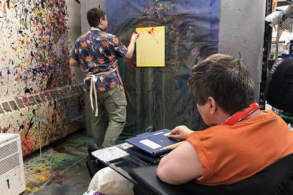 Amy Myers watching as a facilitator throws red paint onto a yellow canvas.