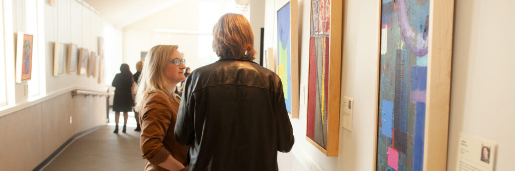 Two able-bodied people looking at paintings in a gallery exhibition at the Robert Schonhorn Arts Center.