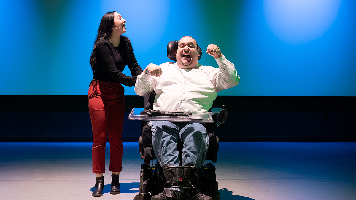 A man in a wheelchair and an able-bodied woman onstage in a theatre.