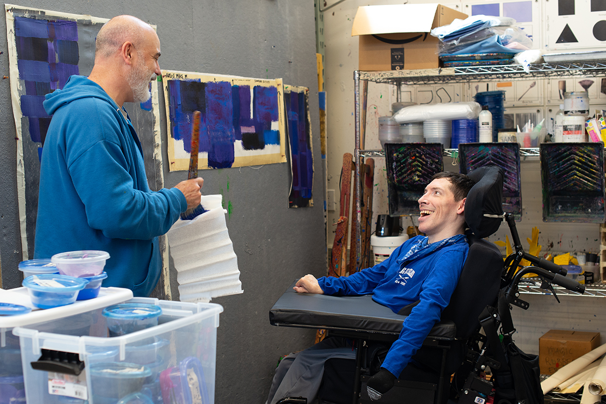 Mike Martin, a man who uses a wheelchair, painting with a facilitator in the Arts Access studio.